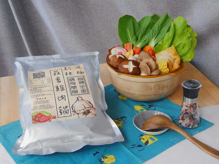 蒜香雞肉鍋銀色真空包放在桌面，旁邊擺有裝滿蔬菜與菇類的砂鍋、胡椒研磨器與木湯匙